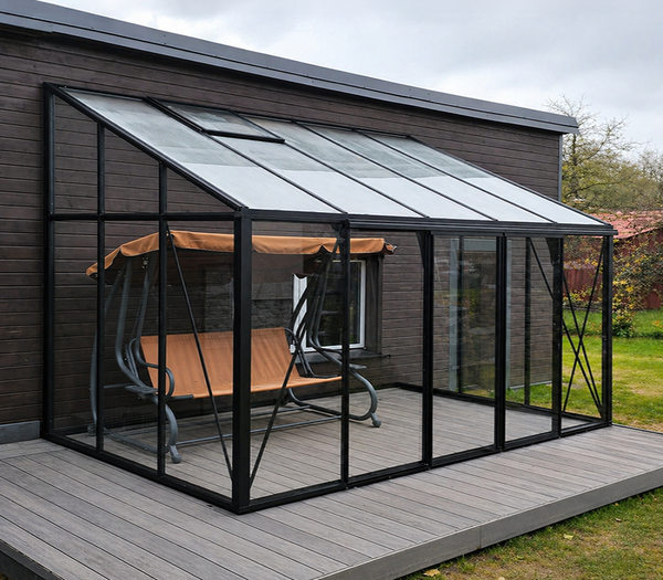 A modern black metal patio sunroom with glass panels on a gray deck, featuring a hanging wooden swing with a tan canopy in front, set against a dark wood house exterior.