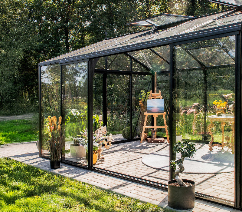 A modern glass greenhouse with a black metal frame, situated outdoors on a paved patio with a lush green garden and trees in the background. Inside, an easel with a painting and potted plants are visible.