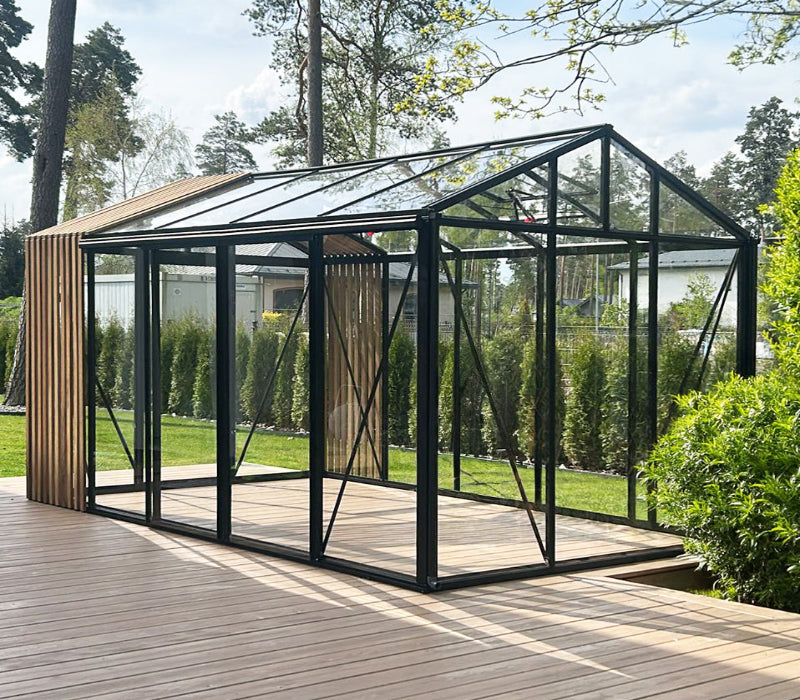 A modern greenhouse with a black metal frame and clear glass panels, situated on a wooden deck in a garden setting.