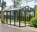A modern greenhouse with a black metal frame and clear glass panels, situated on a wooden deck in a garden setting.