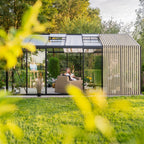 A modern outdoor conservatory with a slatted wood side wall and glass roof, set in a lush green backyard. A woman is relaxing in a wicker sofa inside the conservatory, with an easel and art supplies visible in the background.