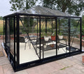 A black-framed glass conservatory with a pitched roof, shown outdoors on a paved patio with garden furniture and potted plants inside. Trees and a field are visible in the background.