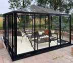 A black-framed glass conservatory with a pitched roof, shown outdoors on a paved patio with garden furniture and potted plants inside. Trees and a field are visible in the background.
