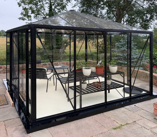 A black-framed glass conservatory with a pitched roof, shown outdoors on a paved patio with garden furniture and potted plants inside. Trees and a field are visible in the background.